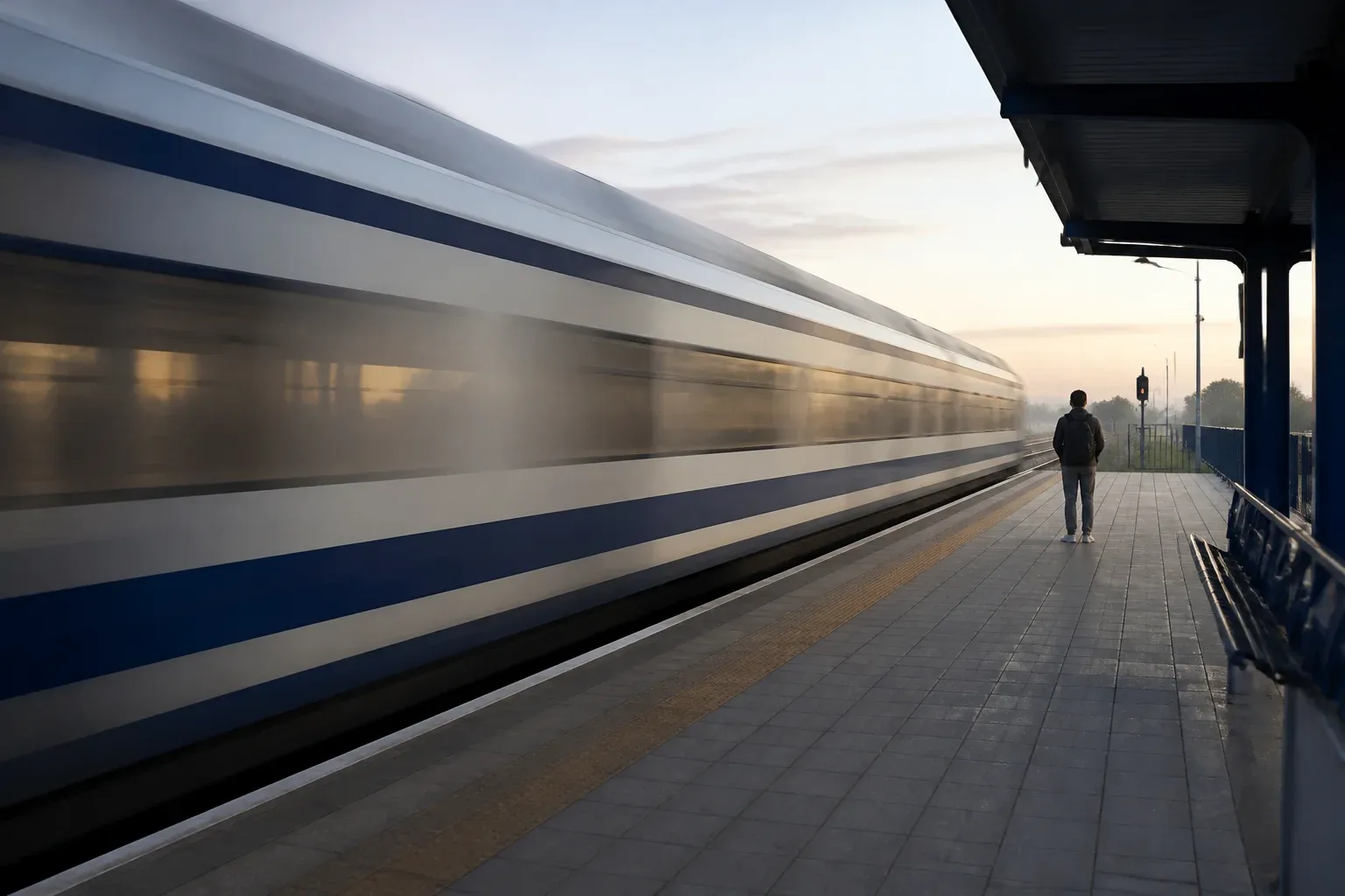 רכבת מהירה חולפת על פני רציף בשעת בוקר, ונוסע בודד ממתין מאחור