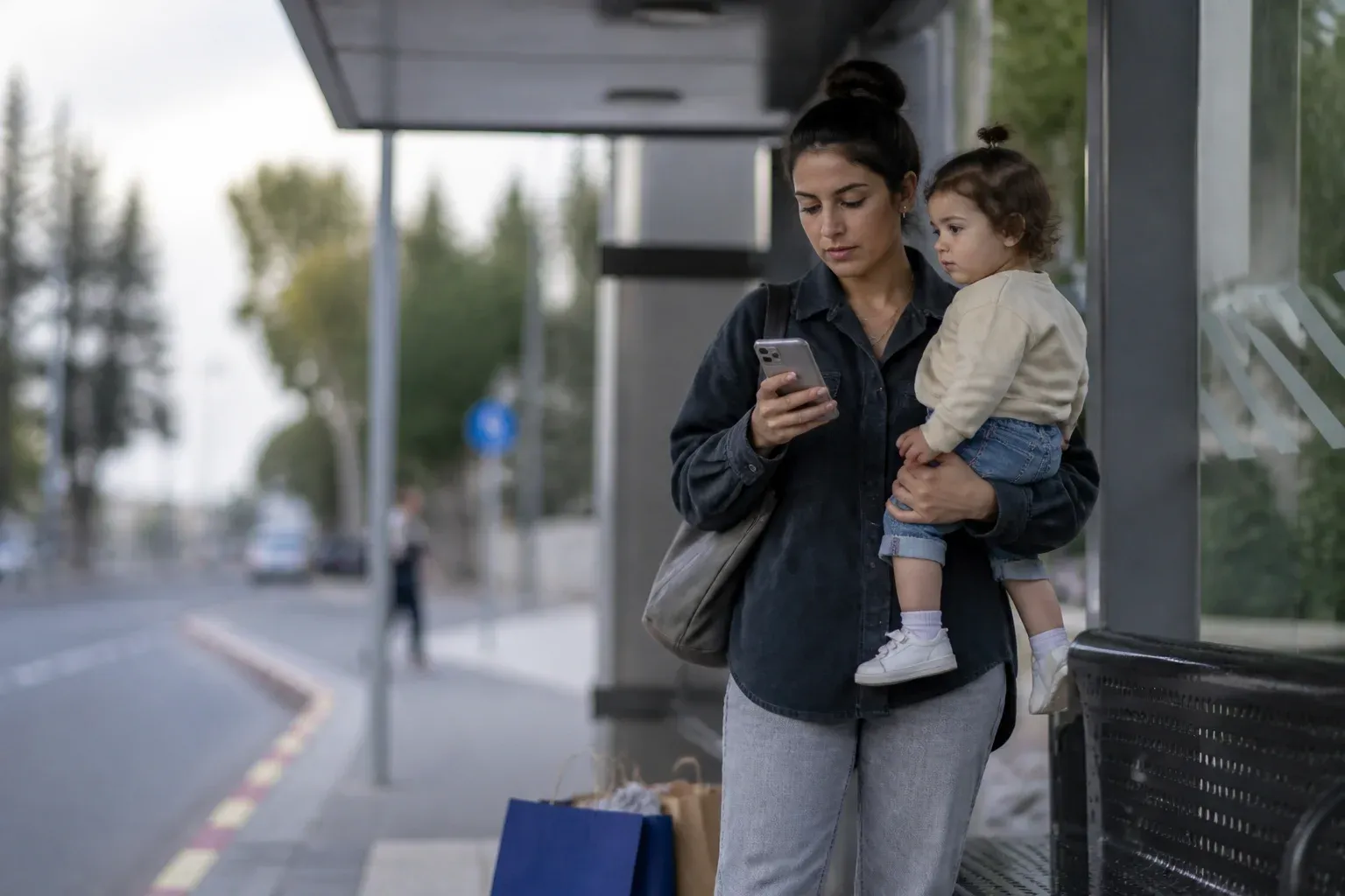 אמא עם פעוט על המותן ממלאת משהו בטלפון בתחנת אוטובוס