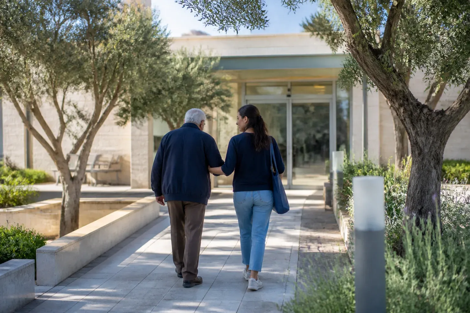 אב מבוגר ובתו הולכים לכניסת זכוכית של קליניקה בין עצים